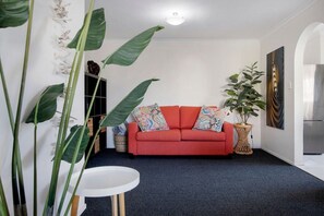 Una sala de estar con un sofá rojo, una mesa blanca redonda y plantas verdes grandes.