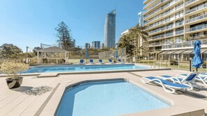 A swimming pool with lounge chairs and a gazebo, surrounded by a fence and palm trees, with a cityscape in the background. - Sunny Surfers Stay - Beach & Tram at your doorstep (Surfers Paradise)