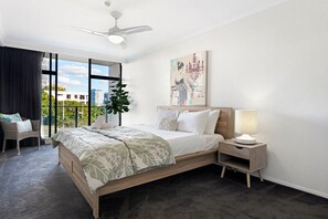 Ein modernes Hotelzimmer mit einem großen Bett, einem Nachttisch, einem Deckenventilator, einem Fenster mit Blick auf Gebäude und einer Zimmerpflanze.