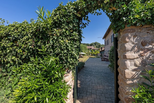 Un chemin de pierres mène à un bâtiment rustique, avec un mur de pierres d’un côté et une haie verdoyante de l’autre.