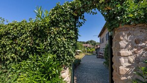 A stone pathway leading to a rustic building with a stone wall on one side and a lush green hedge on the other.