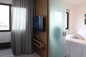 A hotel room with a bed, a TV mounted on a wooden panel, a window with curtains, and a small shelf with decorative items.