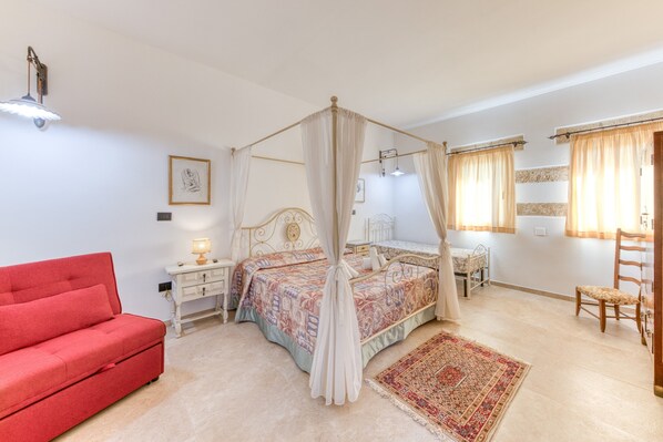 A bedroom with a four-poster bed, a red sofa, a nightstand with a lamp, a wooden chair, and a large rug.
