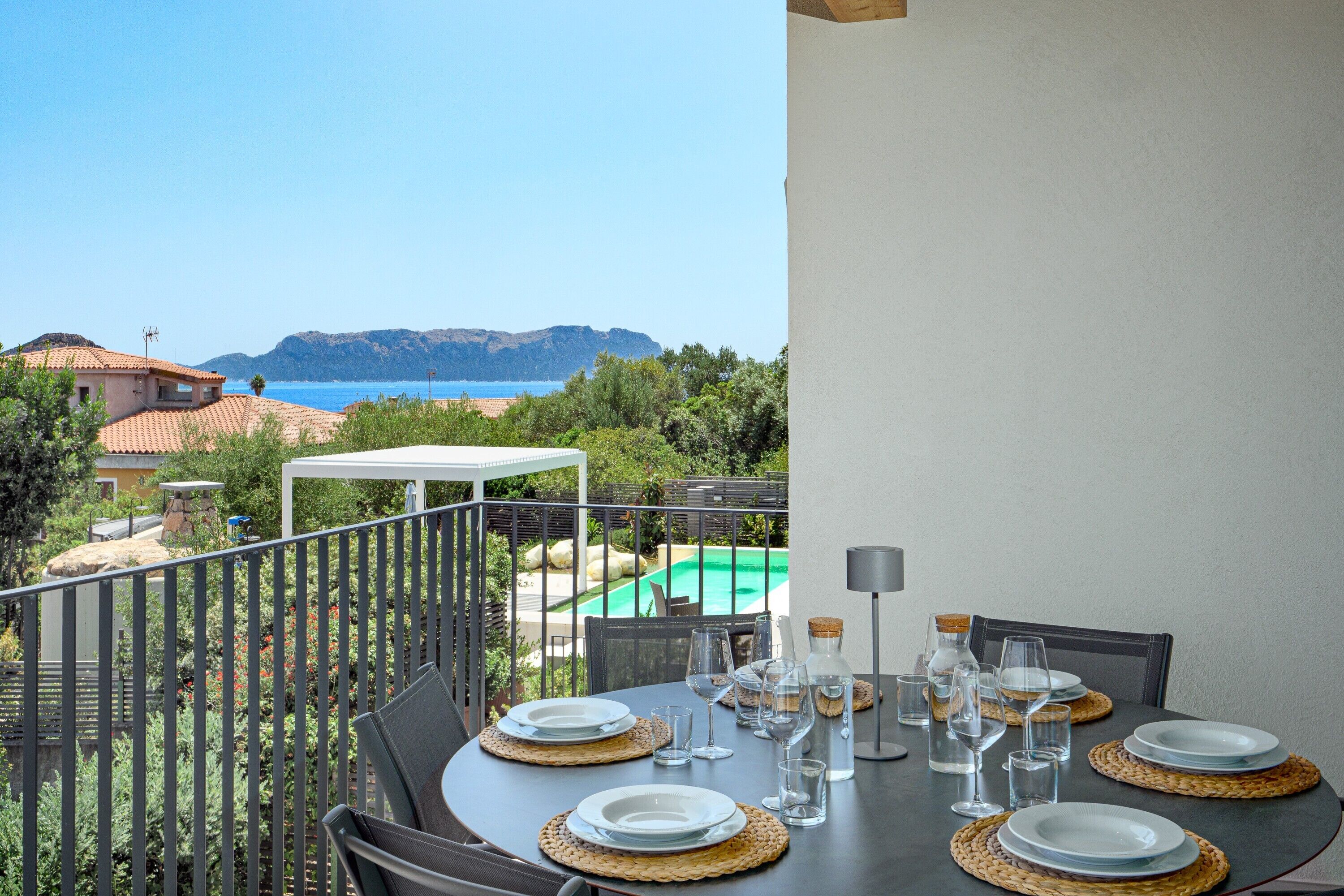 A dining table set for four with plates, glasses, and a candle, overlooking a pool and mountains.