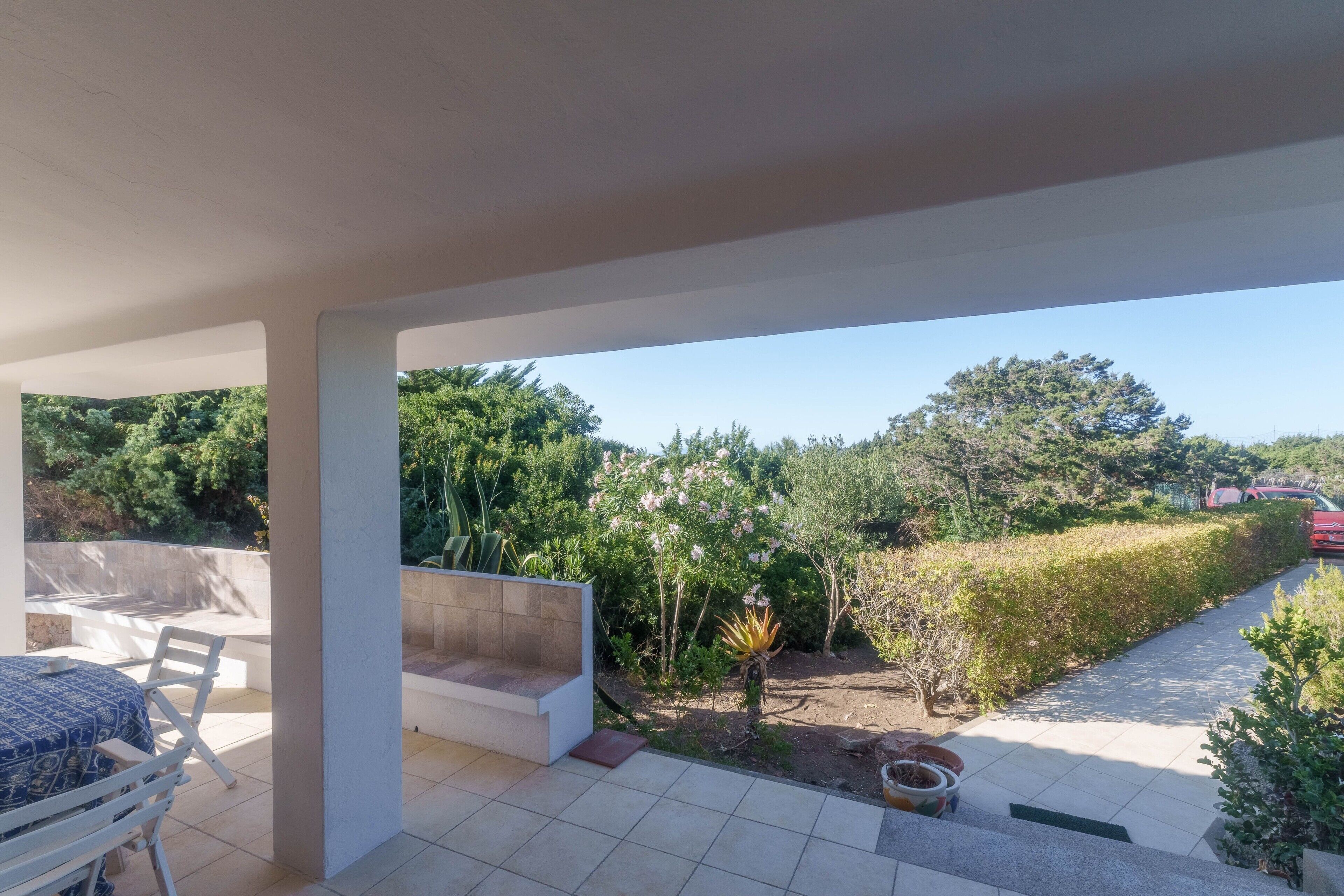 Une terrasse couverte avec une table et des chaises, un petit coin salon et une vue sur un jardin agrémenté d’arbres et d’une haie.
