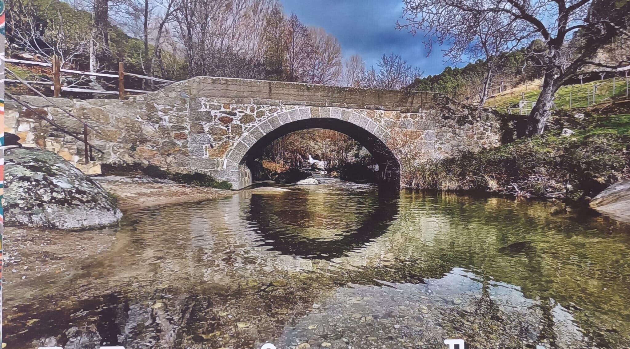 A stone bridge over a clear water body with a rocky shore and trees in the background.