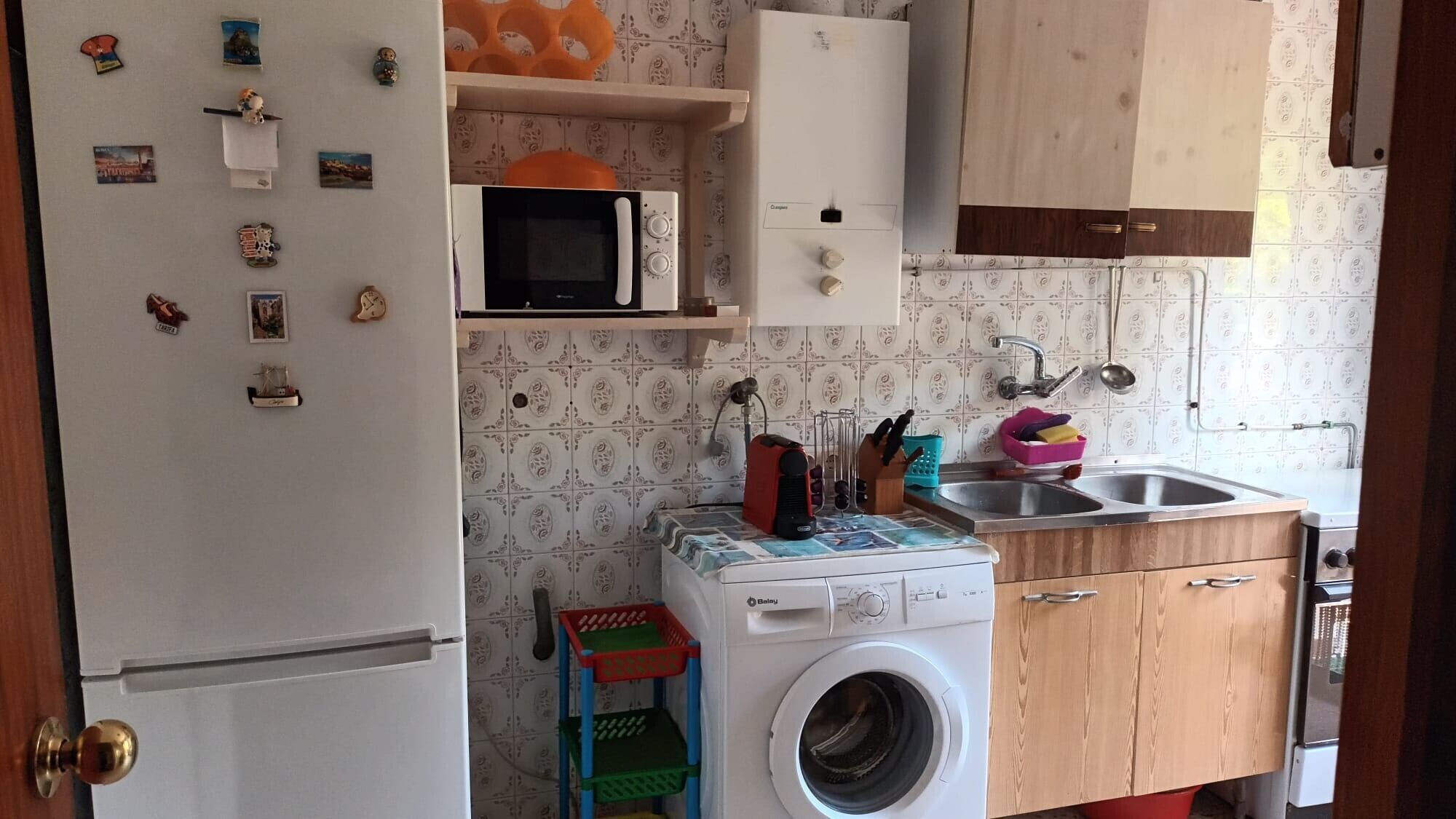 Een compacte keuken met een magnetron, wasmachine en houten kastjes.