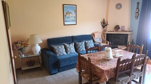 A living room with a blue sofa, patterned cushions, a dining table with a patterned tablecloth, a fireplace, and a framed picture on the wall.