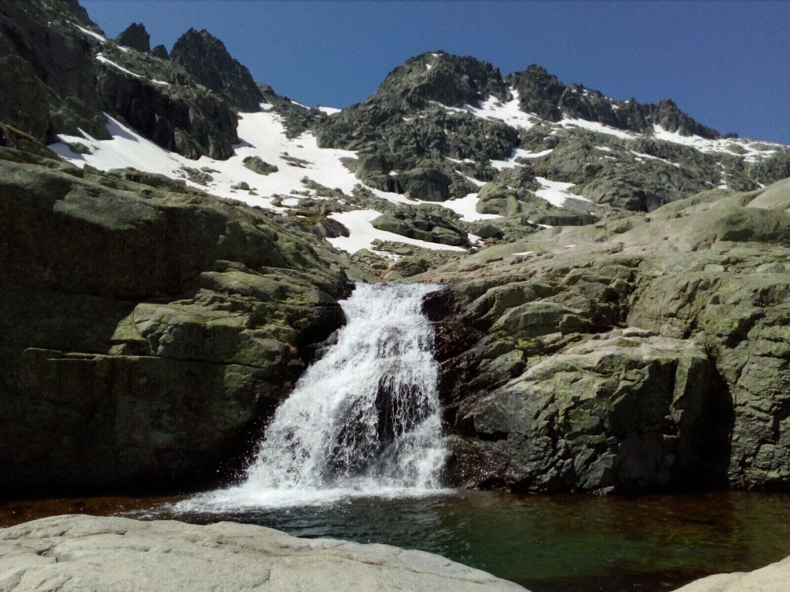 Una cascada en zona montañosa con terreno rocoso y zonas con nieve.