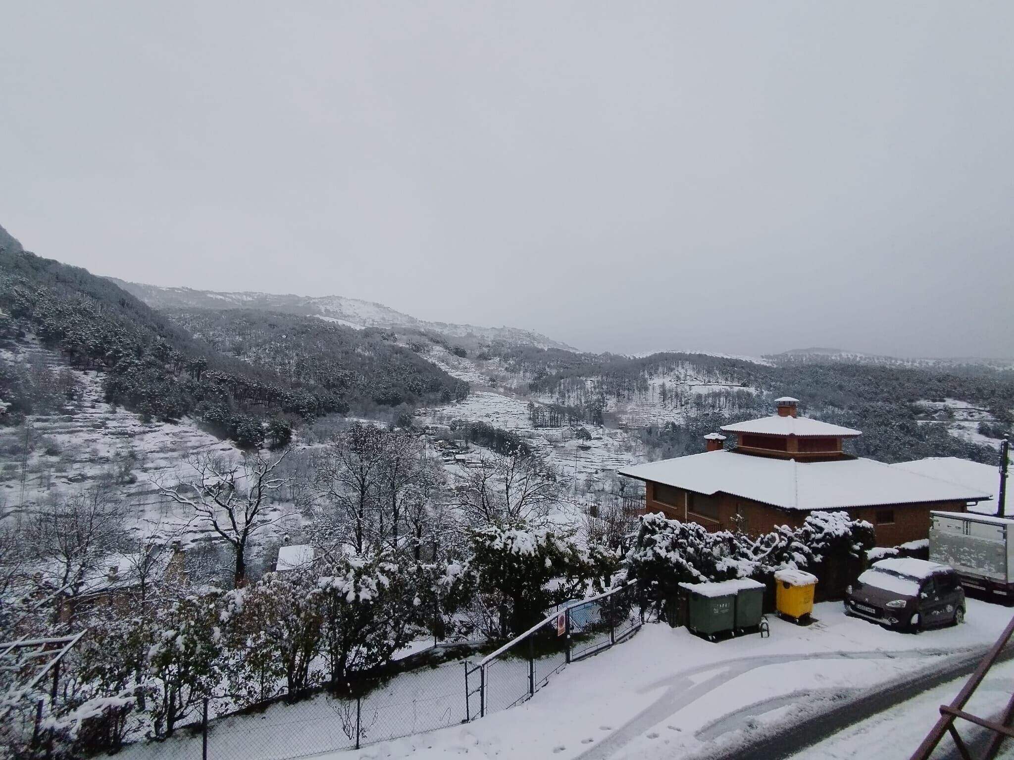A snow-covered landscape with a building that has a traditional roof and a fenced area with a vehicle and bins.
