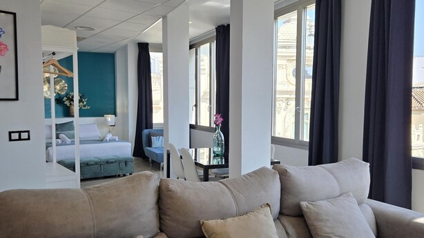 A modern hotel room with a large beige sofa, a bed with a blue headboard, a small table with a vase, and large windows with dark curtains. - Modern Suites in Malaga Center (Málaga)