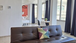Et moderne hotelværelse med en lædersofa, en farverig pude, et træbord og udsigt til en historisk bygning gennem store vinduer.