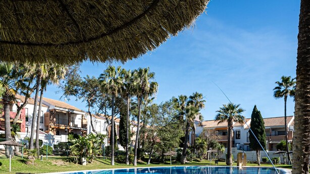 A swimming pool surrounded by palm trees and a building with a tiled roof. - Charming apartment in Chiclana with pool and parking (Chiclana de la Frontera)