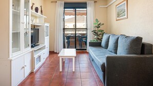 A living room with a grey sofa, a white coffee table, and a TV on a cabinet. There is a window with curtains, a plant, and a picture on the wall. - Charming apartment in Chiclana with pool and parking (Chiclana de la Frontera)