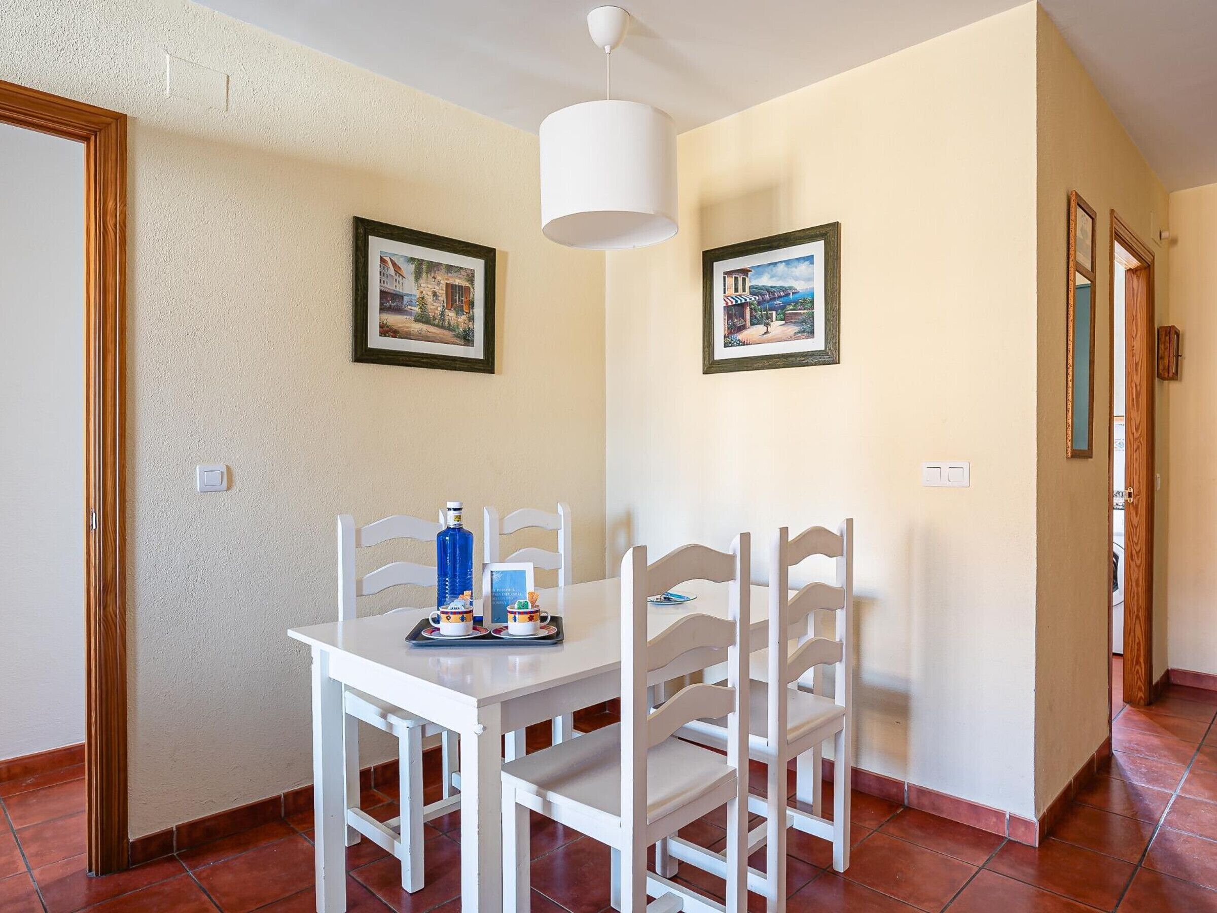 A dining area with a white table and chairs, two framed pictures on the wall, and a blue bottle on the table.