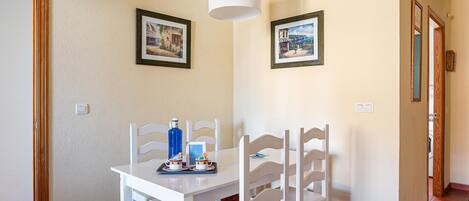 A dining area with a white table and chairs, two framed pictures on the wall, and a blue bottle on the table.