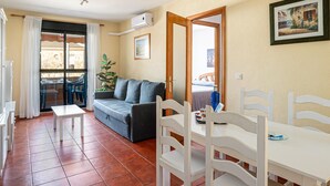 A compact living space with a sofa, dining table, chairs, and a view through a glass door to an outdoor area. - Charming apartment in Chiclana with pool and parking (Chiclana de la Frontera)