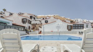 A pool area with white lounge chairs and a small table, surrounded by white buildings with red-tiled roofs.