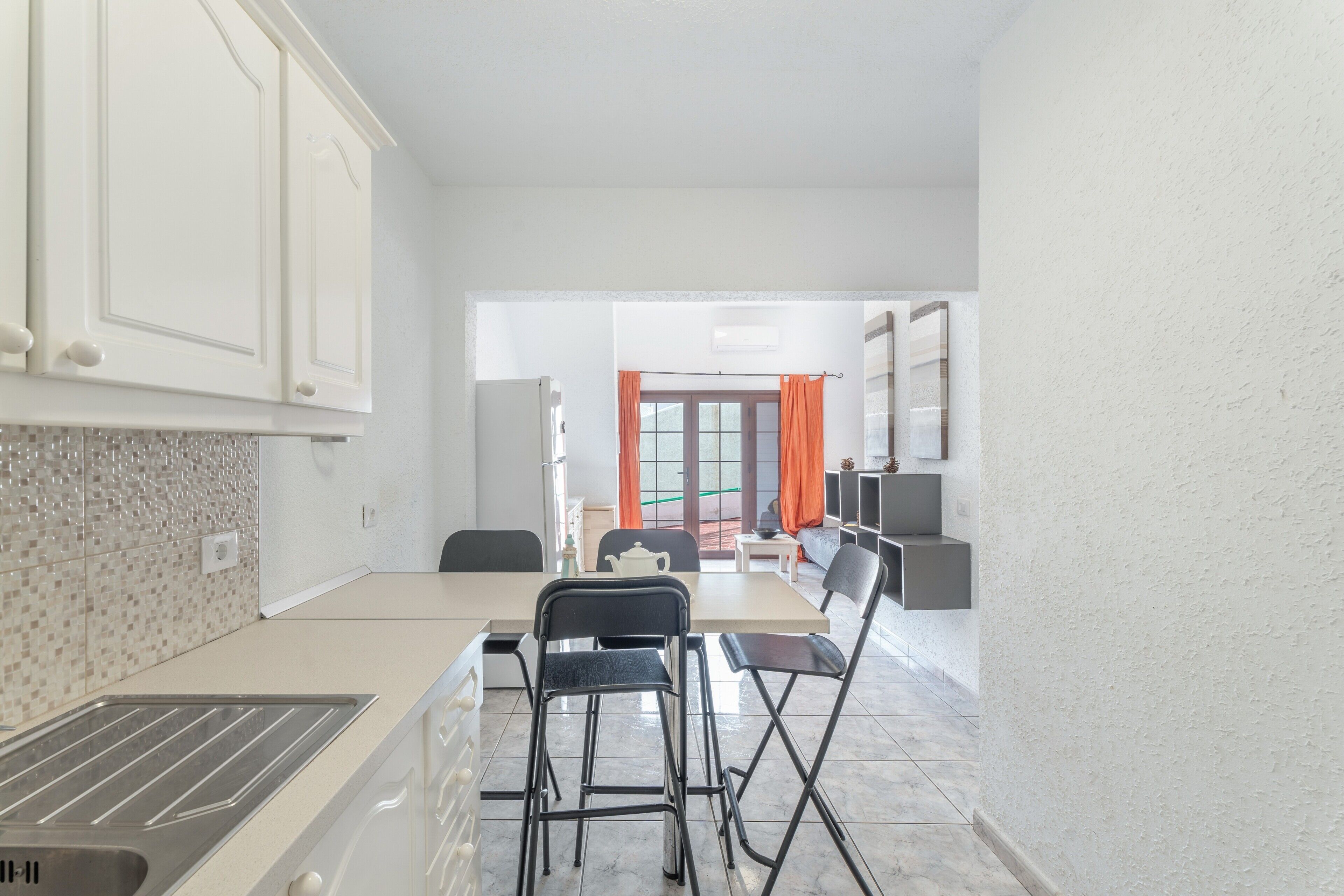 A modern kitchen with a dining area, white cabinets, a sink, and a window with orange curtains.