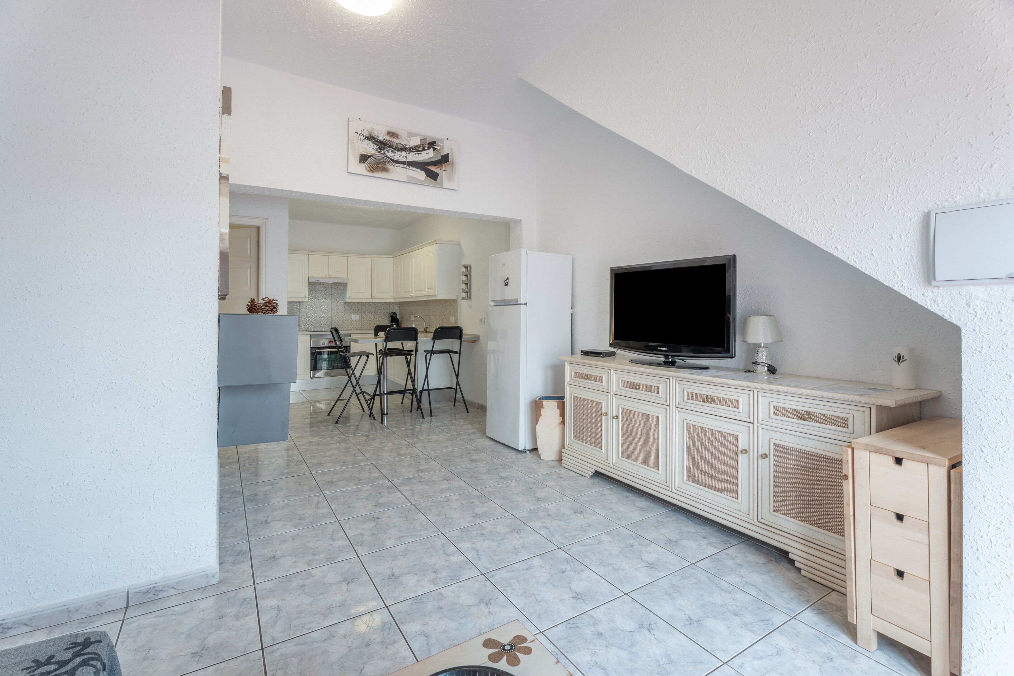 A modern kitchen with white cabinets, a dining area with chairs, a television, and a small cabinet.