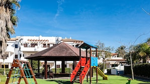 A playground with climbing structures and slides, surrounded by buildings with white facades and red-tiled roofs.