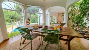 Uma sala de jantar com mesa de madeira, cadeiras verdes e vista para um jardim através de grandes janelas.