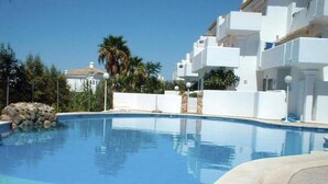 Una piscina azul cristalina rodeada de edificios blancos con balcones, palmeras y un cielo despejado.