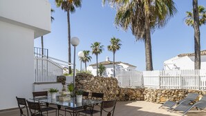 Un patio exterior con una mesa de vidrio y sillas de mimbre, rodeado por un muro de piedra y palmeras.