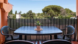 An outdoor patio with a round table and four chairs, a potted plant, and a view of trees and buildings in the background.
