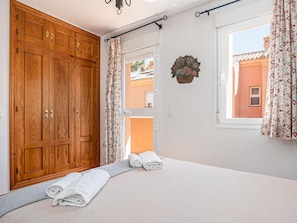 Ein Schlafzimmer mit einem Holzschrank, einem Bett mit weißen Leinen, einem Fenster mit floralem Vorhang und Blick auf ein Gebäude.