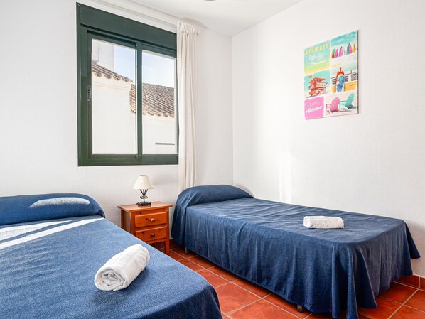 A room with two beds, a window with a view of buildings, a small table with a lamp, and a colorful poster on the wall.