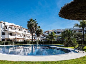 A swimming pool surrounded by a well-maintained lawn, with a bench and a thatched umbrella nearby. - Apartment in Chiclana de la Frontera near La Barrosa Beach (Chiclana de la Frontera)