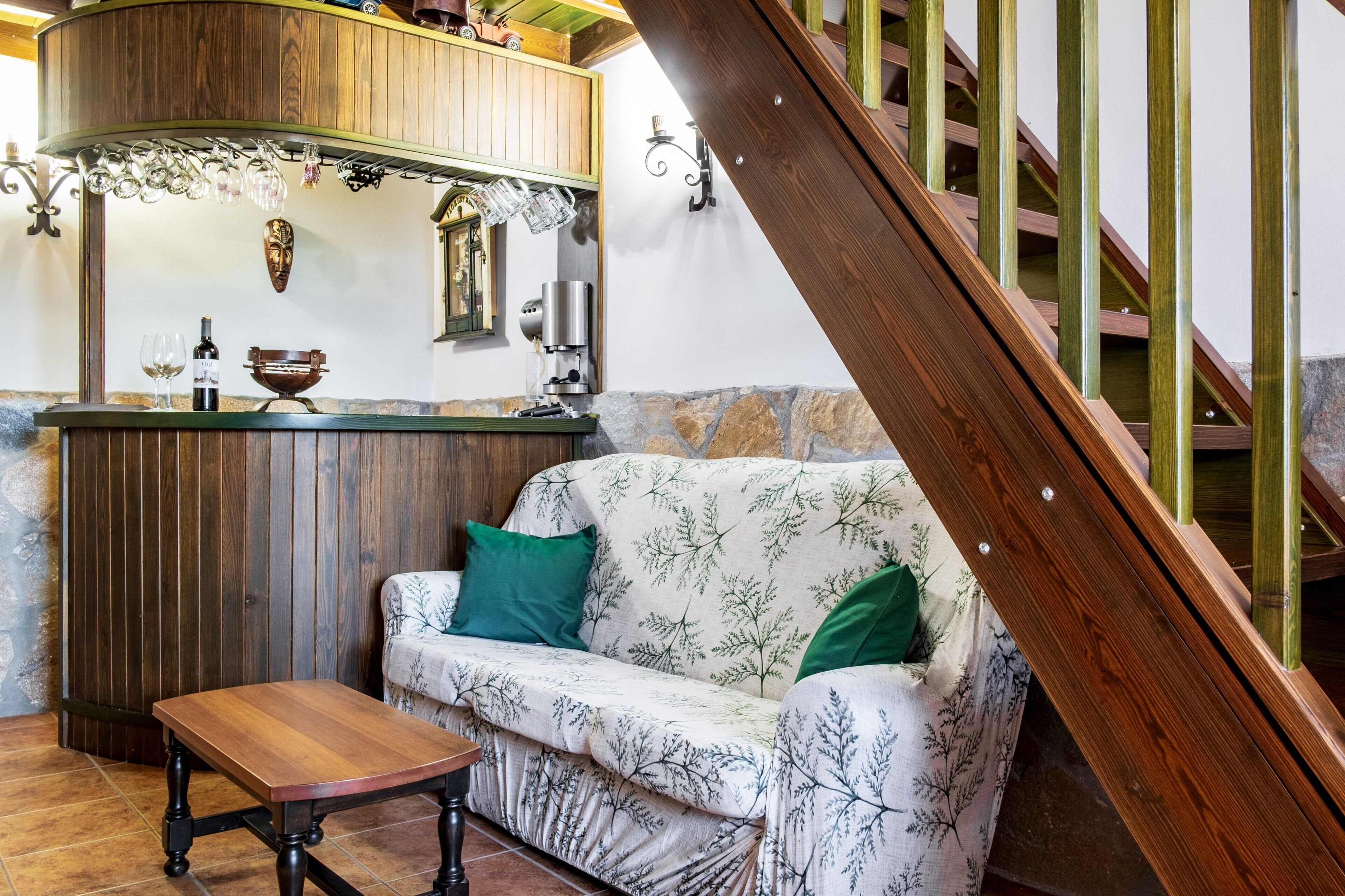 Een houten trap, een houten tafel, een bank met groene kussens en een bar met een fles wijn en glazen.