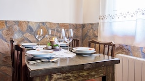 A dining table set with plates, glasses, and cutlery, with a stone wall and a window with a floral curtain in the background.