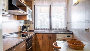 A compact kitchen with wooden cabinets, a built-in oven, a coffee maker, and a window with lace curtains.