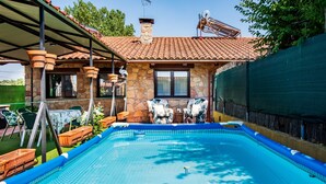 A swimming pool with a stone house in the background, a covered patio, and outdoor furniture.