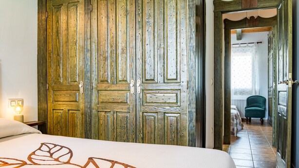 A room with a bed, wooden wardrobe, and a view into another room with a chair and curtains.