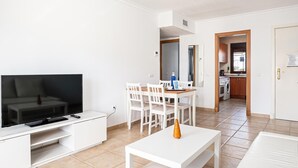 A modern living room with a TV, white furniture, a dining area, and a kitchen visible through an open door. - Holiday Apartment in Chiclana with Air Conditioning and Pools (Chiclana de la Frontera)