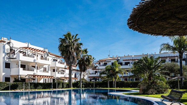 A swimming pool surrounded by a well-maintained lawn, with a bench and a thatched umbrella nearby. - Holiday Apartment in Chiclana with Air Conditioning and Pools (Chiclana de la Frontera)