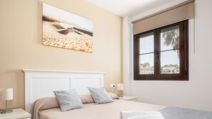 Un dormitorio con cama, almohadas, mesita de noche, lámpara, ventana con vistas y un cuadro en la pared.