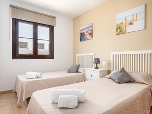 A hotel room with two beds, a window with blinds, a bedside table with a lamp, and a painting on the wall.