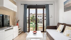 Una sala de estar moderna con un sofá, una mesa de centro, un televisor y un balcón con vistas a edificios y zonas verdes.