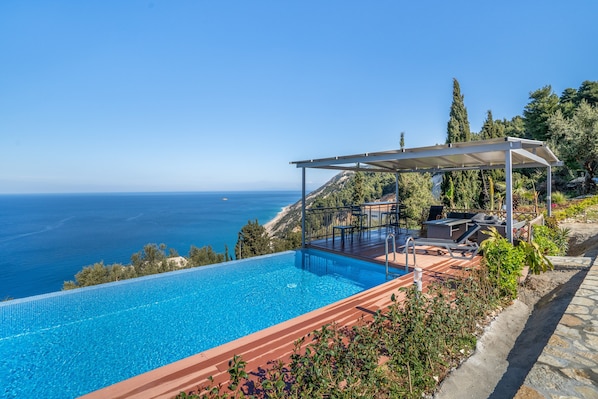 Ein Pool mit Meerblick, ein überdachter Sitzbereich mit Liegestühlen und ein Steinweg.