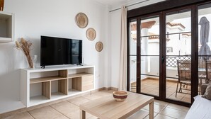 A modern living room with a flat-screen TV on a white console, a wooden table with a bowl, and a sliding glass door leading to an outdoor patio.