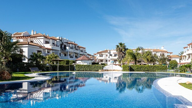 A swimming pool with clear blue water, surrounded by white buildings with red-tiled roofs and balconies.
