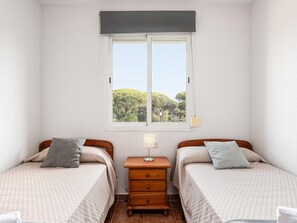 A bedroom with two beds, a window with a view of trees, a bedside table with a lamp, and a wooden dresser.
