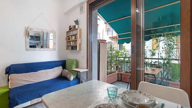 A room with a blue sofa, a small table with a glass and plates, a bookshelf, and a view of the outside balcony. - ‘Terrazzo Del Gelsomino’ Apartment with Private Terrace (Rapallo)