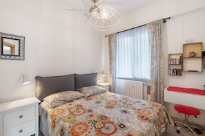 Une chambre à coucher comprenant un lit, une table de chevet, une lampe, une fenêtre avec des rideaux, un radiateur et un bureau avec une chaise.