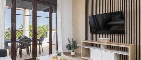 Una sala de estar moderna con un televisor montado en una pared de paneles de madera, un ventanal corredero que da a un espacio exterior, y una mesa de centro de madera con una bandeja.
