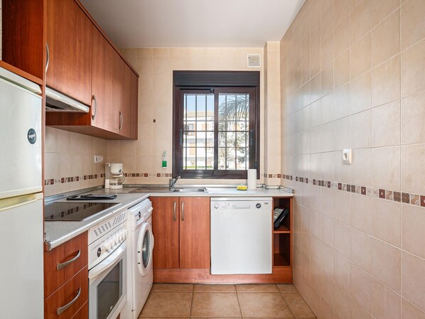 A compact kitchen with wooden cabinets, a built-in oven, a dishwasher, and a window with a view of buildings.
